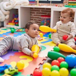 Two adorable babies playing with balls and bowling pin sitting on floor at kindergarten